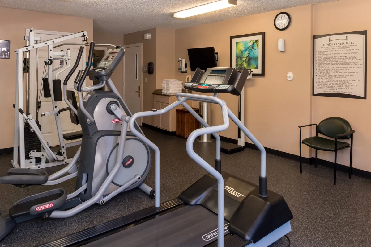 A bright, modern fitness center equipped with elliptical machines, a treadmill, and a weight machine. The space is clean and motivating, with fitness-themed artwork and a visible clock for convenience.