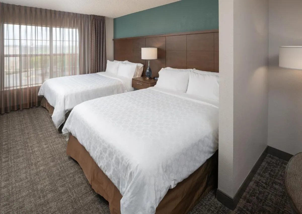 The image showcases a bright and welcoming studio interior featuring two queen beds with fresh linens, a spacious window that bathes the room in natural light, and a cozy nightstand with a lamp. The scene exudes comfort and relaxation, inviting guests to unwind and recharge.