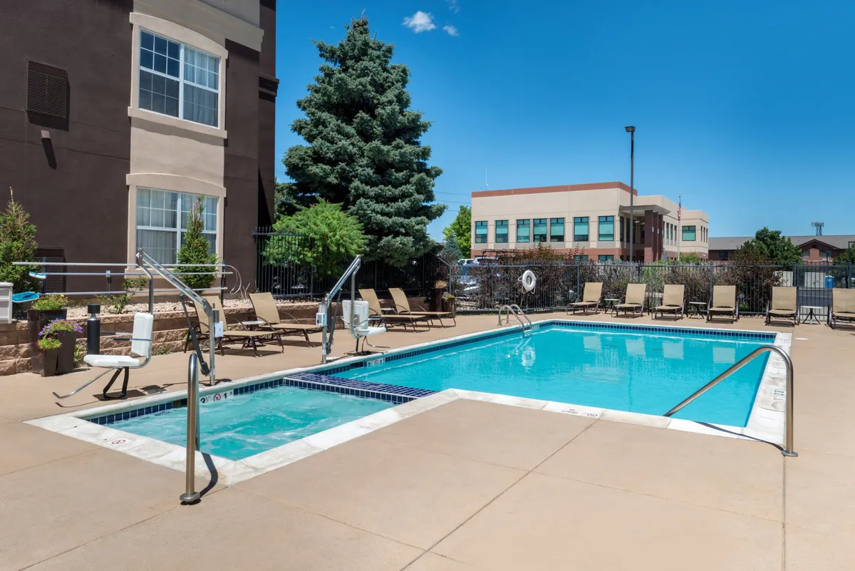 An inviting exterior scene featuring a vibrant swimming pool surrounded by lounge chairs and lush landscaping. People are seen enjoying the pool and relaxing poolside, highlighting the resort-style atmosphere.