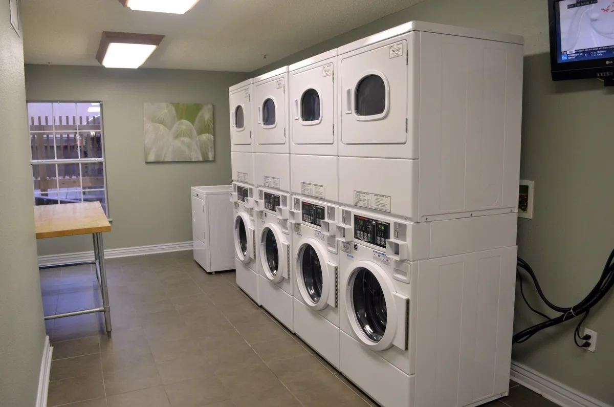 This image features a bright, practical laundry room with stacked washing machines and dryers, a counter table for folding, and a window that lets in natural light. The space is clean, organized, and showcases the on-site laundry amenities available to guests.