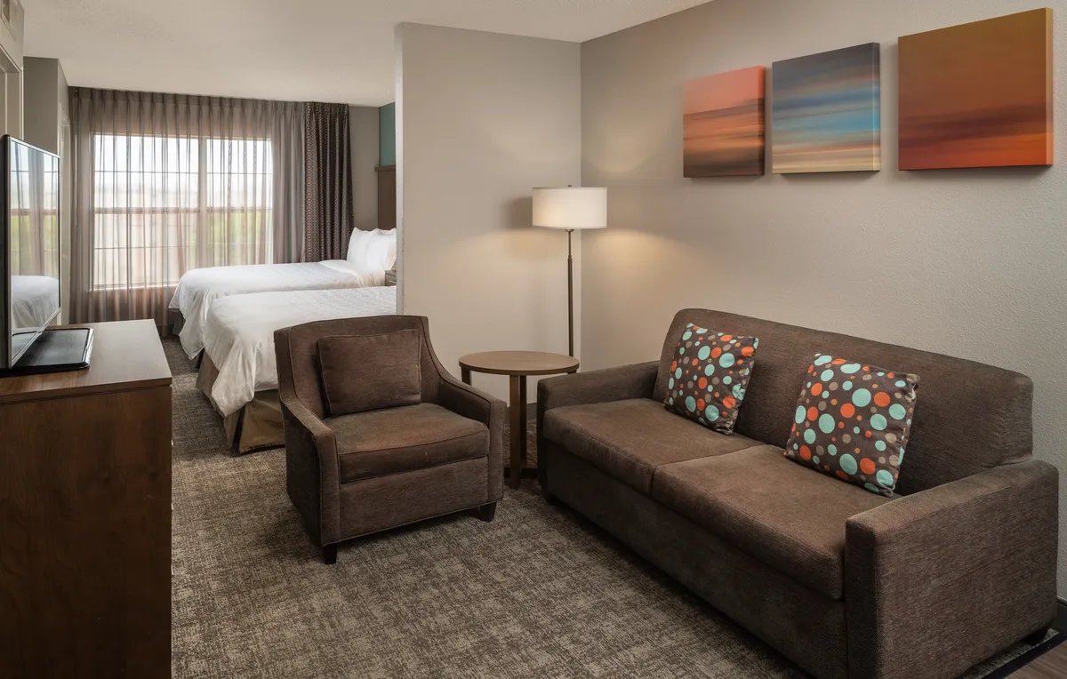 A cozy studio space featuring a sofa with colorful cushions, vibrant artwork on the wall, and a bed visible in the background. The layout seamlessly blends the living and sleeping areas, offering a welcoming atmosphere that feels personalized and comfortable.