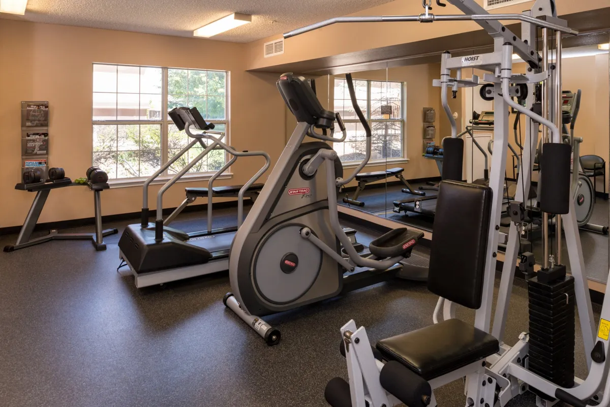 The image showcases a well-equipped fitness center featuring an elliptical machine, weight machine, and exercise bike, set against a mirrored wall. The space is bright and organized, inviting both solo workouts and group fitness activities.